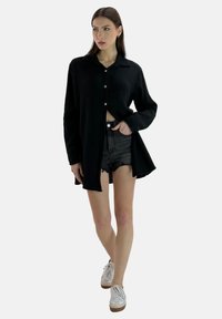 Sort langærmet skjorte med knapper lavet af struktureret stof, kombineret med slidte sorte shorts og hvide sneakers med brune såler.