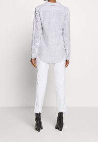 Person som bär en vit långärmad skjorta med svarta vertikala ränder, vita byxor och svarta klackade ankelstövlar, stående framför en enkel bakgrund.