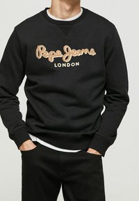 Hombre vistiendo sudadera negra con el logo beige de "Pepe Jeans London", combinada con pantalones negros y una camisa blanca debajo. Manos en los bolsillos.
