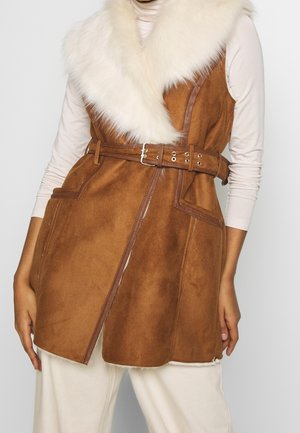 Braune Wildlederweste ohne Ärmel mit weißem Kunstpelzkragen, tailliert mit Gürtel, getragen über einem hellbeigen Langarmshirt und cremefarbenen Hosen.