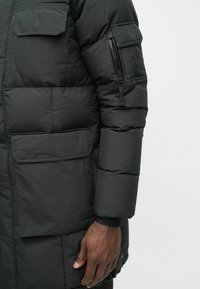 Črn pufast plašč z žepom na zip na rokavu, debelo quilted teksturo in flap žepom spredaj. Ima logotip na rokavu.