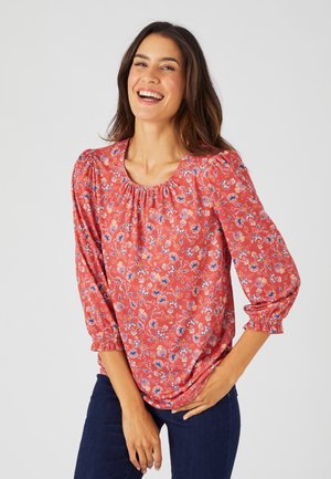 Rode bloes met een ronde halslijn, geplooide details en driekwartmouwen. De stof heeft kleine blauwe en witte bloemenpatronen.