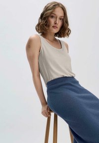 Hellbeige Tanktop mit taillierter Passform, kombiniert mit einem langen, dunkelblauen, strukturierten Rock. Model sitzt auf einem Holzstuhl vor einem schlichten Hintergrund.