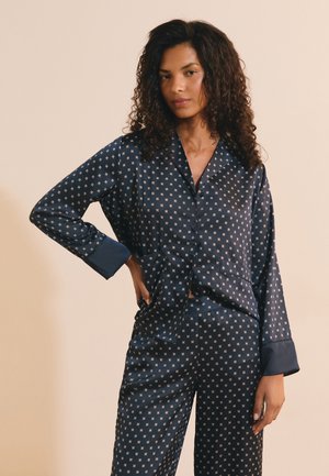 Donna con capelli ricci che indossa un completo pigiama in raso blu navy con leggero motivo geometrico, in piedi con una mano sui fianchi su uno sfondo uniforme.