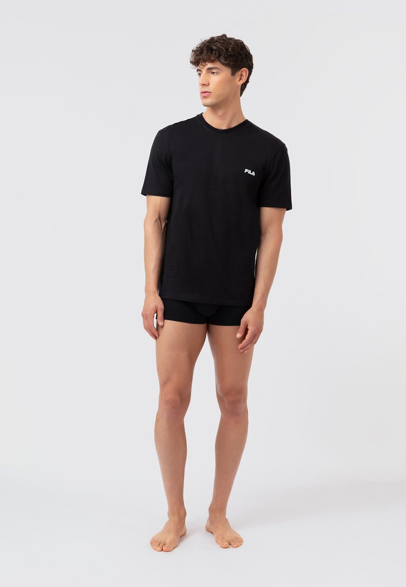 Νέος άνδρας, ξυπόλητος, φοράει μαύρο t-shirt FILA και μαύρο σορτς, κοιτάζει αριστερά σε απλό φωτεινό φόντο.