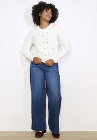 Bílý, huňatý cardigan s kulatým výstřihem a dekorativními stříbrnými ozdobami, zkombinovaný s rozšířenými modrými džíny a vínovými teniskami.