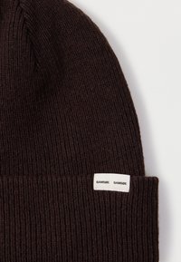 Gorro de punto marrón con textura acanalada. Etiqueta blanca con la marca "SAMSØE" cosida en el borde inferior. Material suave y cálido.