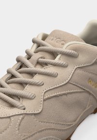 Baskets en daim beige avec de grosses lacets beige, coutures apparentes, et logo "HUGO" partiel doré sur la panneau latéral.