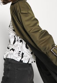 Giacca lunga satin verde oliva, indossata sopra una maglietta bianca con un motivo astratto di mani e volti neri. Abbinata a jeans grigi scuri.