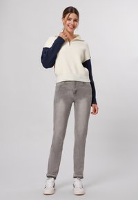 Maglione zip-up bianco e blu navy con maniche a coste, abbinato a jeans grigio chiaro e sneakers bianche. Texture liscia, design informale.