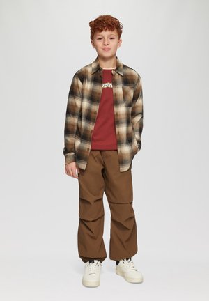 Ein rothaariger Junge steht da und trägt ein braun kariertes Hemd über einem roten Grafik-T-Shirt, braune Hosen und weiße Turnschuhe vor einem einfarbigen Hintergrund.