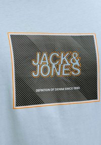 Lyseblå skjorte med et grafikpatch med teksten "JACK & JONES" i fed orange, omgivet af en stribet sort baggrund og hvid tekst nedenunder.