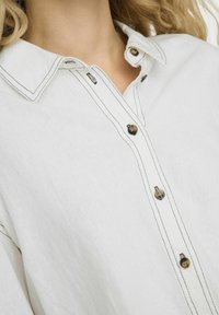 Biała lniana koszula z ostrym kołnierzykiem, z przodu zapinana na ciemne brązowe guziki, z kontrastowym przeszyciem i luźnym krojem.