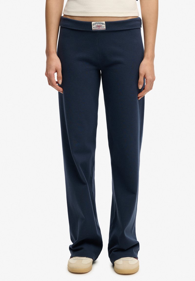 Persona con pantalones azul marino de corte recto con una etiqueta visible de Superdry en la cintura y zapatillas beige, de pie quieta.