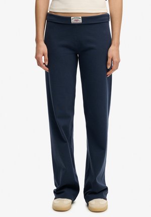 Person wearing navy blue straight-leg pants with a visible Superdry label at the waistband and beige sneakers, standing still.