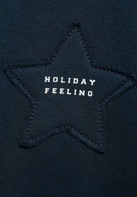 Námornícky textil s hvězdným nášivkou a bílým stehem, na němž je v tučném písmu vyveden nápis "PRÁZDNINOVÝ POCIT".