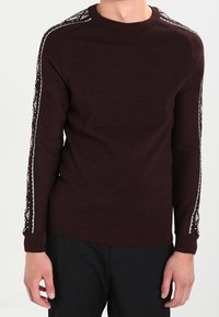 Burgundy sweater with a round neckline, ribbed cuffs, and contrasting patterned accents on sleeves in red and white. Soft knit texture.