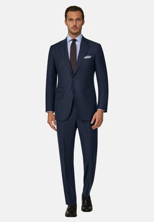 Homme debout portant un costume bleu foncé à deux boutons avec une chemise bleu clair, une cravate sombre à motifs, une pochette blanche et des chaussures habillées noires.