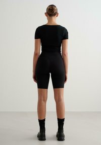 T-shirt nero aderente e pantaloni corti neri in tessuto morbido, indossati con calze sportive nere e scarpe nere ingombranti, visti da dietro.