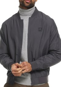 Grijze bomberjack van gladde stof met een ritssluiting aan de voorkant, geribbelde manchetten en een klein embleem op de borst. Draag het over een lichtgrijze coltrui.