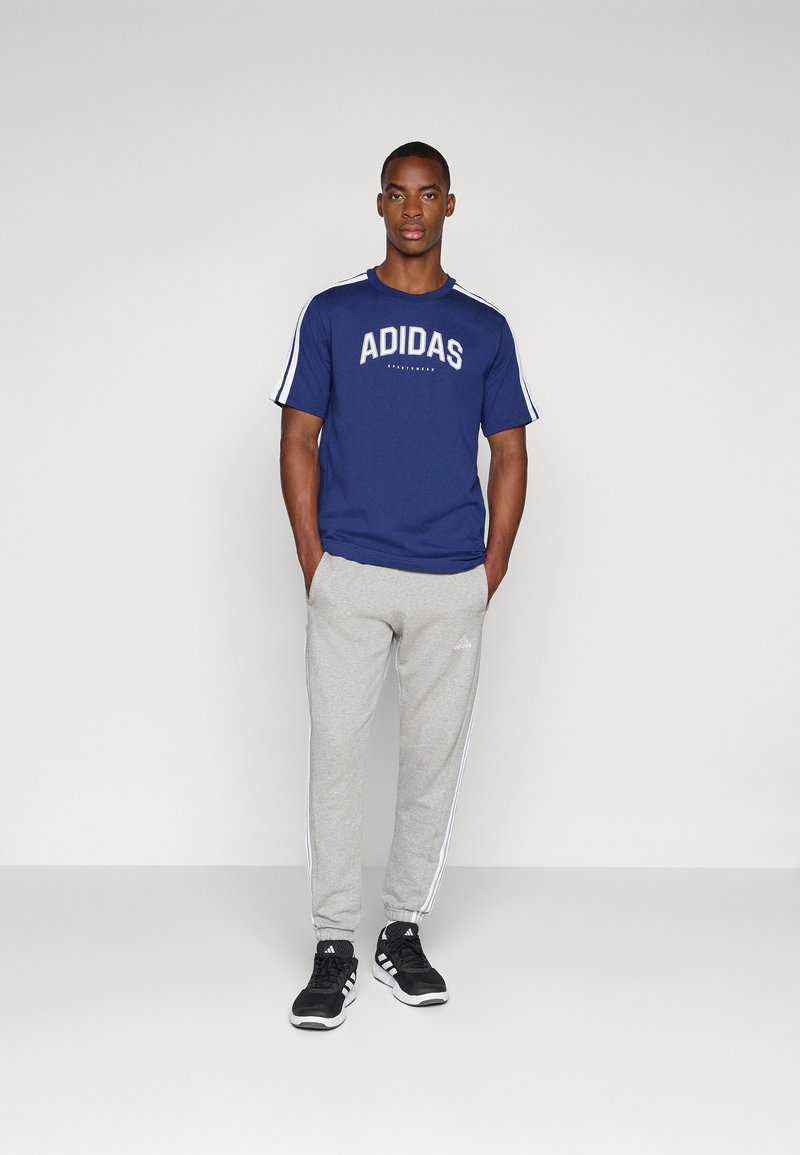 Granatowa koszulka adidas z białym logo, szare dresy z białymi bocznymi paskami i czarne buty sportowe. Prosty, codzienny strój sportowy.
