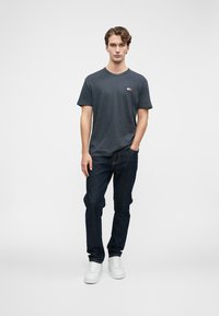 Mladý muž stojící s jednou rukou v kapse, oblečený v tmavě šedé košili s krátkým rukávem, tmavých džínách a bílých teniskách na bílém pozadí.