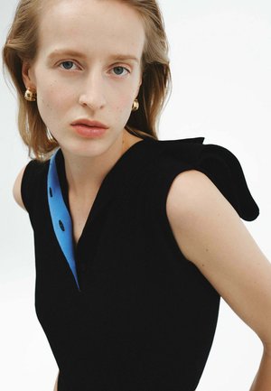 Vestido negro con cuello azul, detalles de botones y acentos de hombros oversized. La modelo lleva pendientes dorados y el cabello lacio.