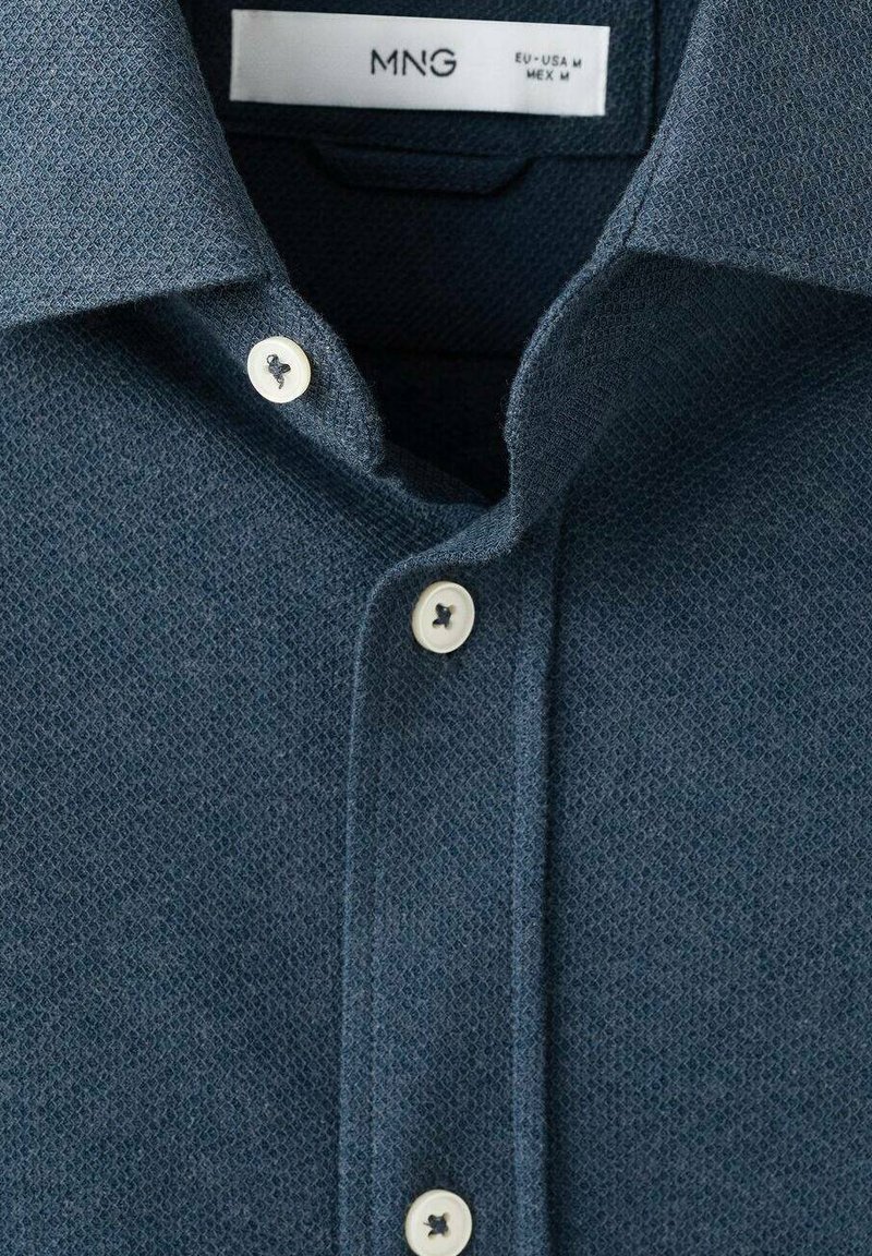 Mørkeblå tekstureret skjorte med krave, der kan knappes ned, prydet med to hvide knapper og et vævet mønster. Plejeetiket synlig indeni kraven.