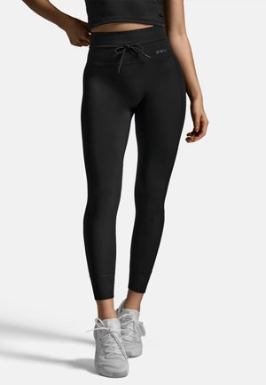Schwarze High-Waist-Leggings mit Kordelzug, figurnahem Schnitt, glatter Textur und Logo-Detail. Kombiniert mit weißen Sportschuhen.