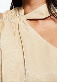 Haut beige avec un design en tissu froncé, texture satiné, et un large col avec un accent noué à l'épaule.