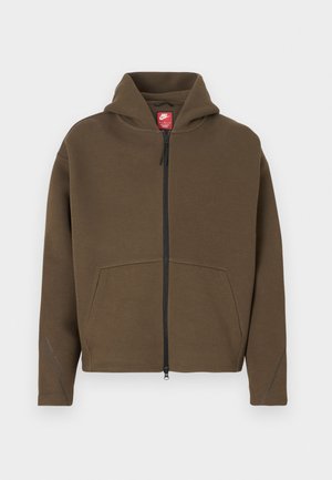 Καφέ φούτερ με φερμουάρ και μεγάλο κουκούλι, εμπρόσθιες τσέπες και αντανακλαστικές λεπτομέρειες στα μανίκια. Κατασκευασμένο από μαλακό ύφασμα με υφή.