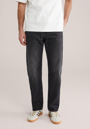 Vīrietis valkā izbalējušas melnas taisnas džinsa bikses, baltus sporta apavus ar brūnām zolēm un ielocītu baltu T-kreklu, stāvot pret neitrālu fonu.