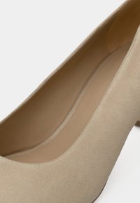 Chaussure à talon haut en daim beige avec un bout arrondi et une doublure intérieure lisse, présentant un design minimaliste et une texture douce.