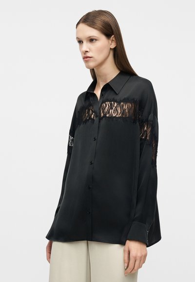 Γυναίκα που φοράει μια υπερμεγέθη μαύρη μπλούζα με κουμπιά και δαντελένιες ενθέσεις στο στήθος και τα μανίκια, συνδυασμένη με μπεζ παντελόνι, σε απλό φόντο.