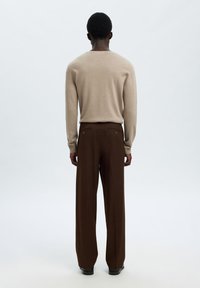 Άτομο που στέκεται γυρισμένο με την πλάτη φορώντας μπεζ μακρυμάνικη μπλούζα, καφέ φαρδιά παντελόνια και μαύρα παπούτσια σε απλό λευκό φόντο.