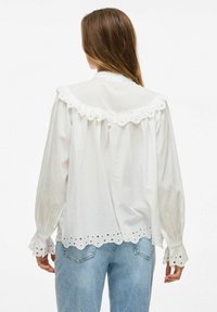 Biała bluzka z długim rękawem z falbaniastego kołnierza i mankietów, z ozdobnymi dziurkami wzdłuż brzegu i mankietów. Wykonana z lekkiego materiału.