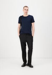 Jūras spēku crew-neck t-kreklis no kokvilnas, apvienots ar melniem pielāgotajiem biksēm un melnām alaizītām kurpēm. Modelis stāv ar rokām kabatās, valkājot krustboda somu.