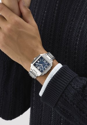 Csuklón ezüst téglalap alakú karóra kék számlappal és római számokkal, fekete kötött pulóverhez és fehér ingmandzsetthoz párosítva.