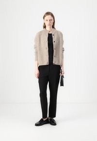 Beige kötött kardigán lekerekített gallérral és fekete gombokkal, fekete toppal és fekete nadrággal, valamint fekete cipővel viselve.
