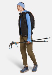 Chaleco aislante negro, camiseta de manga larga azul, pantalones verde olivo, zapatos de senderismo azules, sosteniendo bastones de trekking con empuñaduras de corcho, llevando una cinta para la cabeza azul.