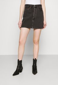 Zwarte denim mini-jurk met een nauwsluitend design, zichtbare voorzakken en stiksels, gecombineerd met zwarte enkellaarsjes.