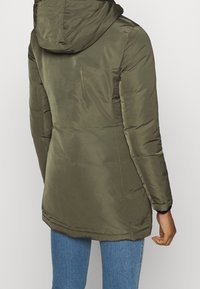 Oliwkowo-zielona pikowana kurtka z kapturem, wykonana z gładkiego, błyszczącego materiału, o długim kroju sięgającym poniżej bioder.