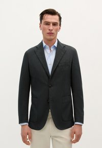 Blazer de cuadros oscuros con solapas de muesca, dos bolsillos frontales y dos botones negros. Llevado sobre una camisa de color azul claro.