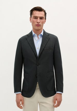 Blazer de cuadros oscuros con solapas de muesca, dos bolsillos frontales y dos botones negros. Llevado sobre una camisa de color azul claro.