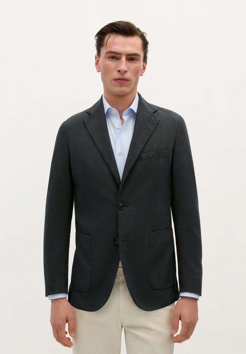 Blazer de cuadros oscuros con solapas de muesca, dos bolsillos frontales y dos botones negros. Llevado sobre una camisa de color azul claro.