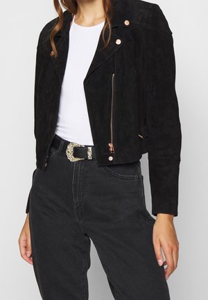 Frau trägt eine schwarze, kurz geschnittene Wildlederjacke mit goldenem Reißverschluss, weißes T-Shirt, hoch taillierte schwarze Jeans und einen schwarzen Gürtel mit verzierter Schnalle.