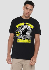 Black cotton t-shirt featuring a large graphic of a dinosaur skull, green leaves, and the text "ROAR-SOME GRANDAD" in yellow and white.