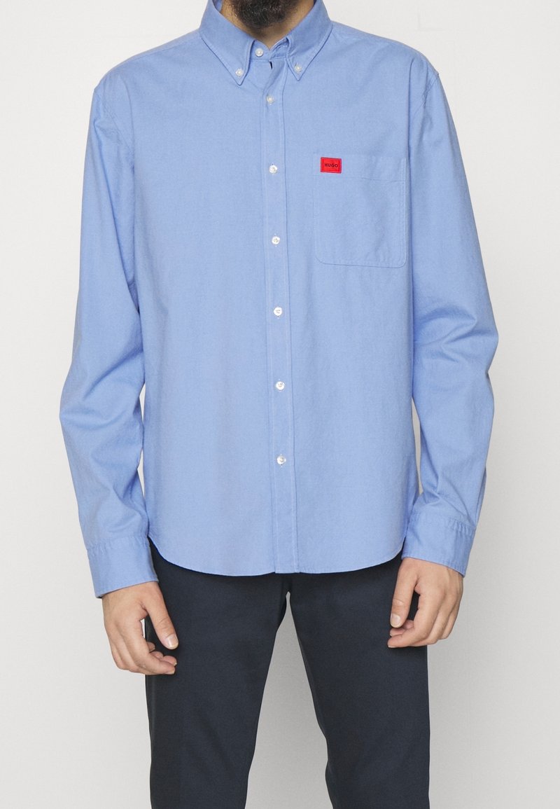 Homme portant une chemise bleu clair boutonnée avec un petit patch logo rouge sur la poche poitrine et un pantalon foncé, debout devant un fond uni.