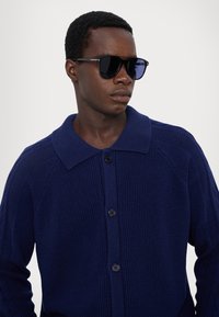 Navy blue knitted cardigan with a wide collar, four buttons, and textured fabric. Black sunglasses with blue-tinted lenses.