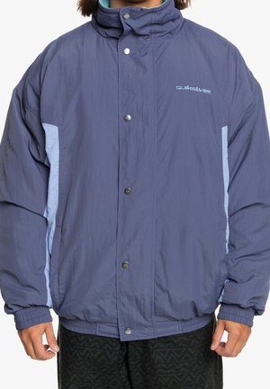 Person trägt eine blaue und hellblaue Quiksilver-Jacke mit Druckknöpfen und elastischen Bündchen, steht vor einem einfarbigen Hintergrund.